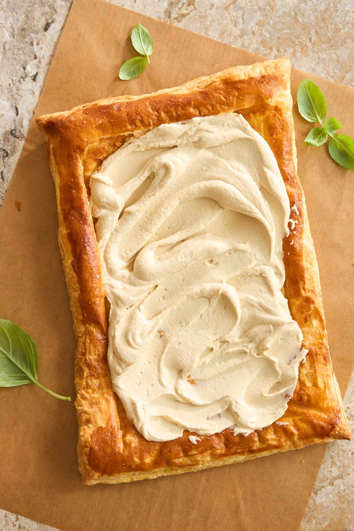 A rectangular sheet of golden-brown puff pastry with a thick layer of creamy spread on top, garnished with a few fresh green basil leaves on the side, on brown parchment paper.
