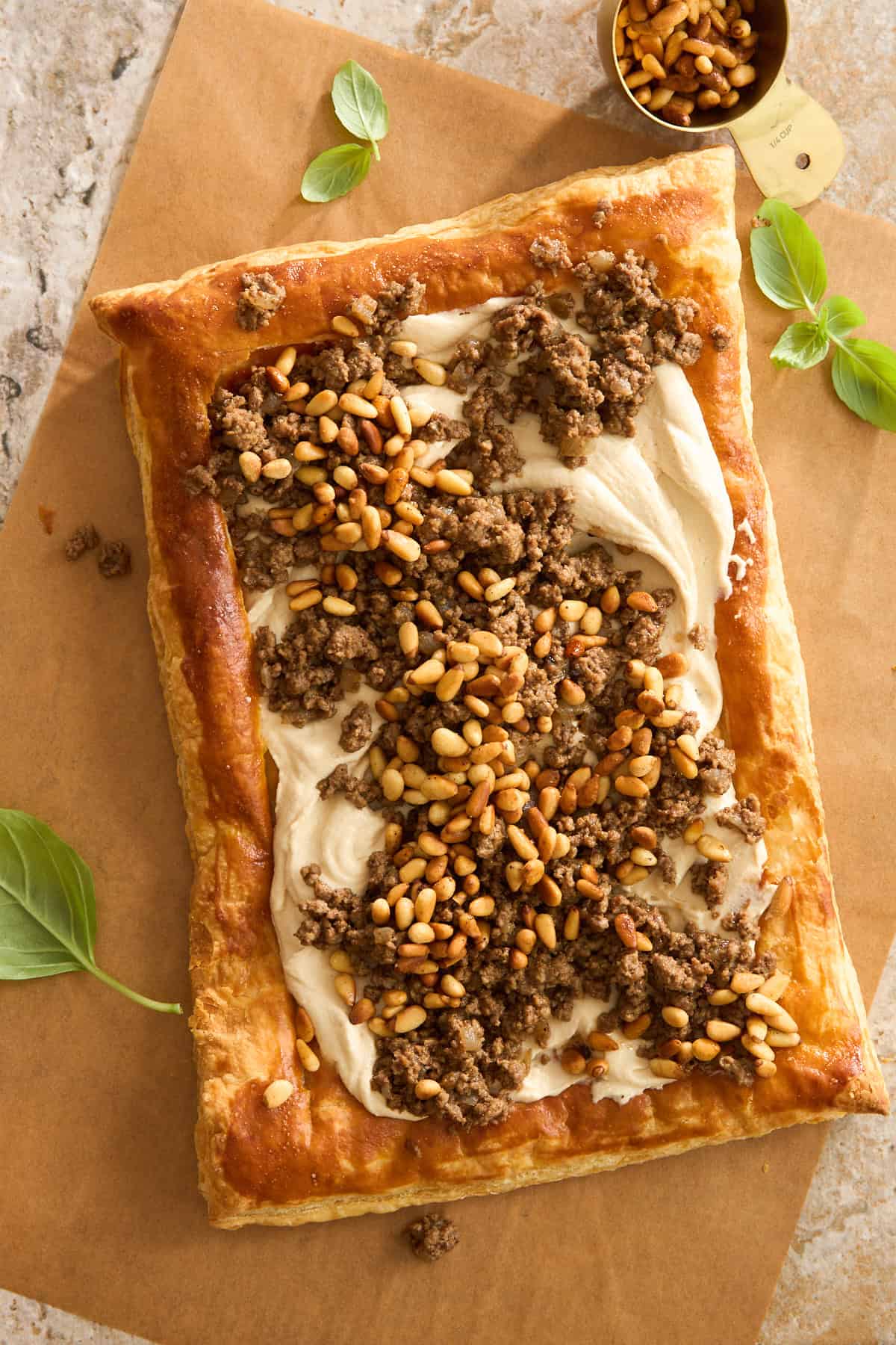 A rectangular puff pastry tart topped with creamy spread, seasoned ground meat, and toasted pine nuts, garnished with fresh basil leaves on parchment paper.