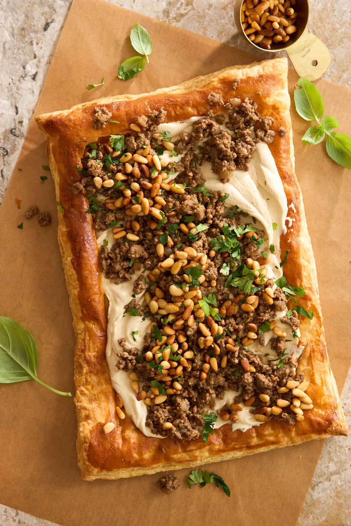 A rectangular puff pastry topped with creamy hummus, cooked ground beef, toasted pine nuts, and chopped parsley sits on parchment paper with basil leaves and a small cup of pine nuts nearby.