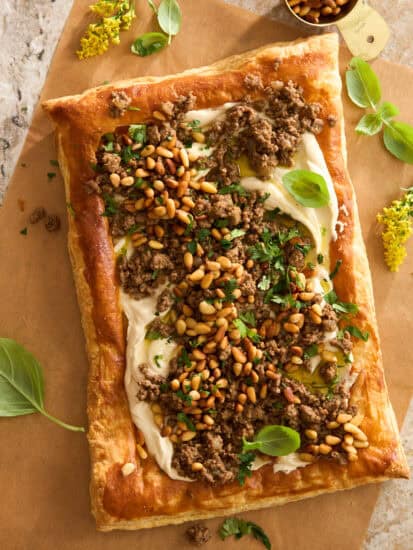 A rectangular puff pastry tart topped with creamy spread, cooked ground meat, toasted pine nuts, fresh herbs, and basil leaves, placed on parchment paper with a gold measuring cup and yellow flowers nearby.