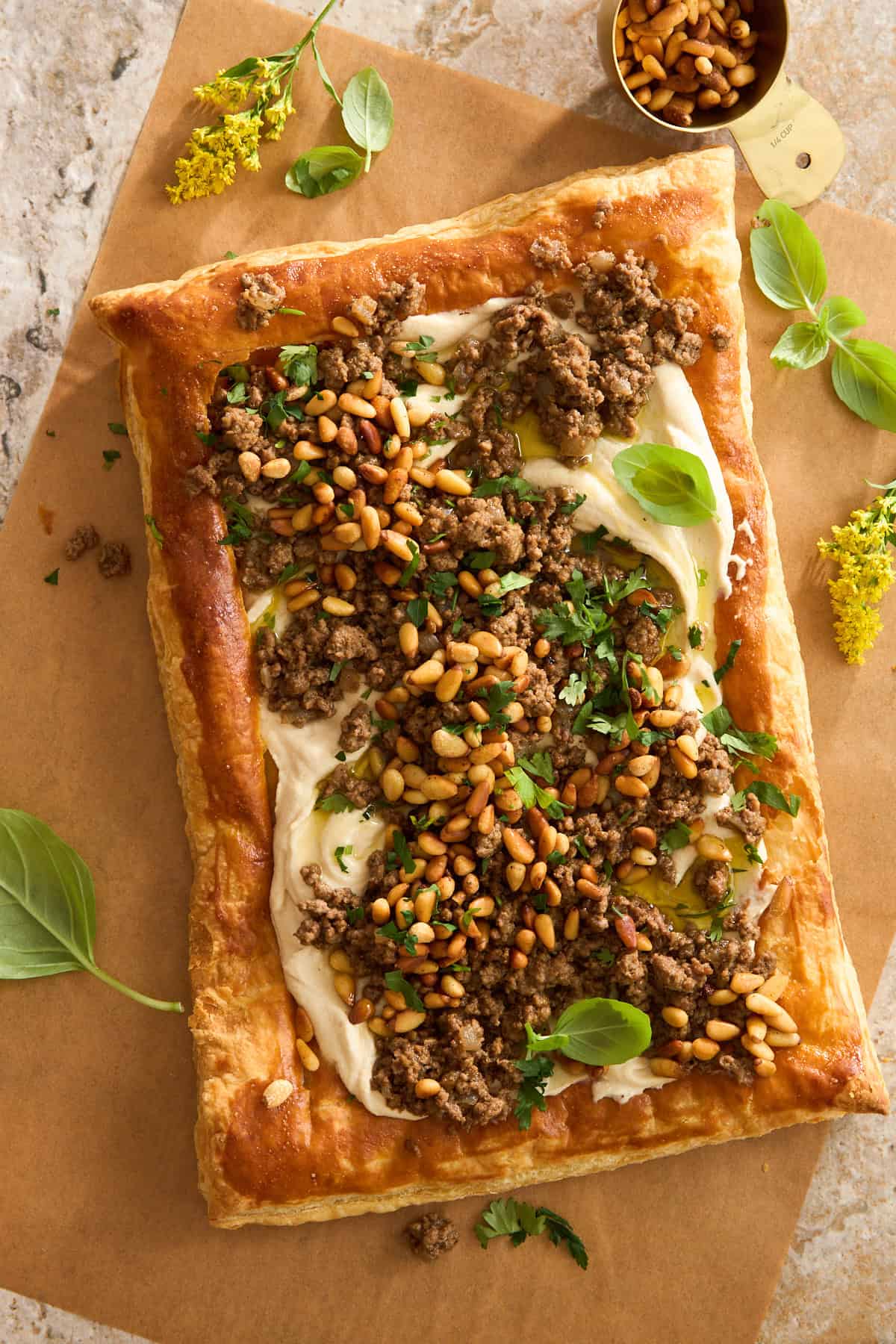 A rectangular puff pastry tart topped with creamy spread, cooked ground meat, toasted pine nuts, fresh herbs, and basil leaves, placed on parchment paper with a gold measuring cup and yellow flowers nearby.