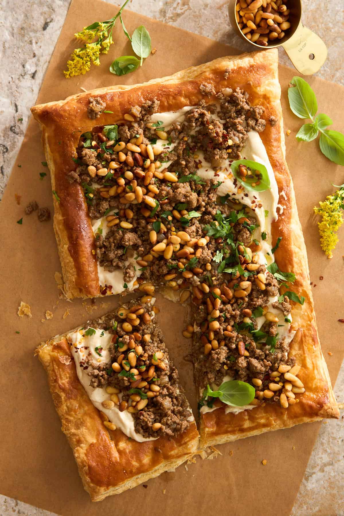 A rectangular puff pastry topped with ground meat, pine nuts, fresh herbs, and a creamy white sauce, partially cut into pieces and arranged on brown parchment paper with scattered yellow flowers and basil leaves nearby.