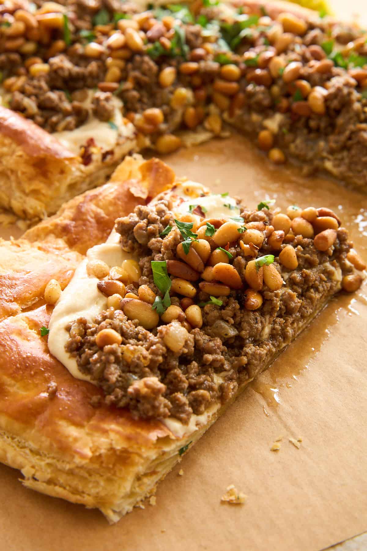 A close-up of a rectangular pastry topped with seasoned ground meat, toasted pine nuts, and chopped parsley, resting on parchment paper. One piece is cut and slightly pulled away from the rest.