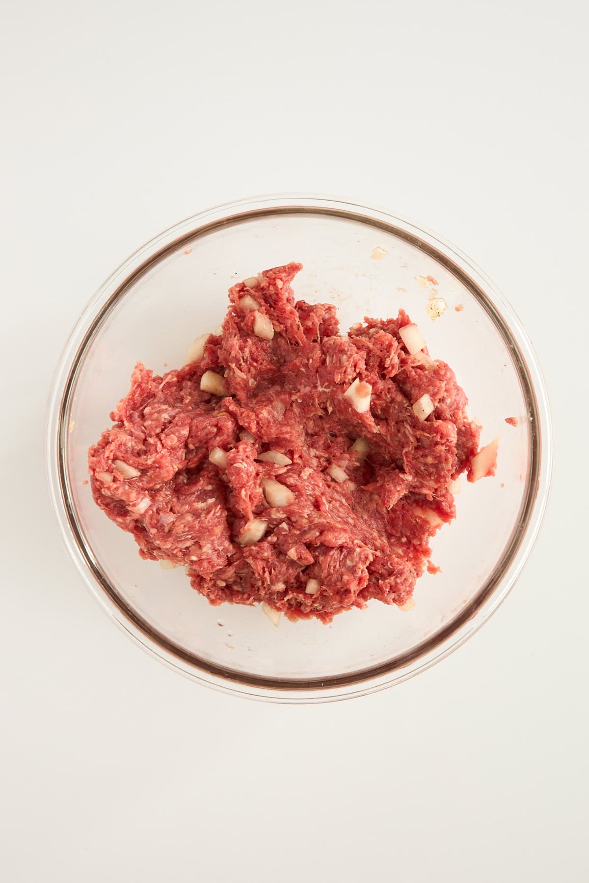 A glass bowl filled with raw ground meat mixed with chopped onions, placed on a white surface.