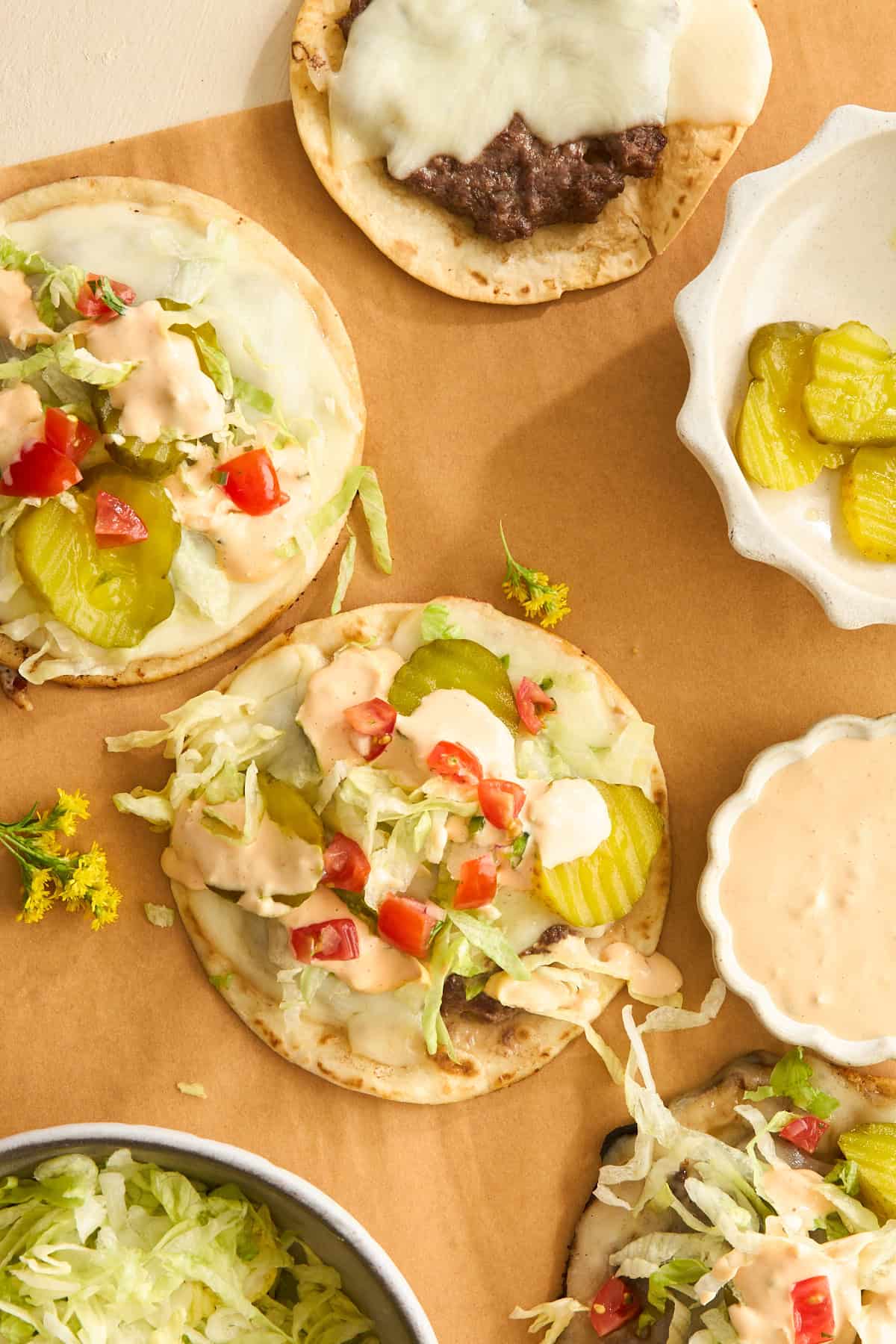 Overhead view of tostadas topped with shredded lettuce, pickles, diced tomatoes, sauce, and melted cheese. A bowl of lettuce, a small dish of pickles, and a bowl of creamy sauce are nearby on a brown surface.