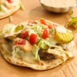 A flatbread topped with ground beef, melted cheese, shredded lettuce, diced tomatoes, pickles, and a creamy sauce sits on brown parchment paper, with a small dish of extra sauce in the background.