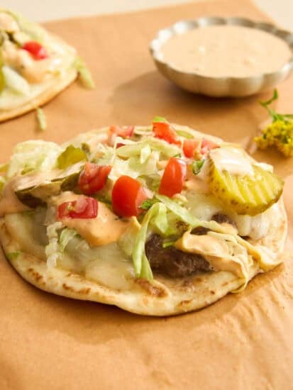 A flatbread topped with ground beef, melted cheese, shredded lettuce, diced tomatoes, pickles, and a creamy sauce sits on brown parchment paper, with a small dish of extra sauce in the background.