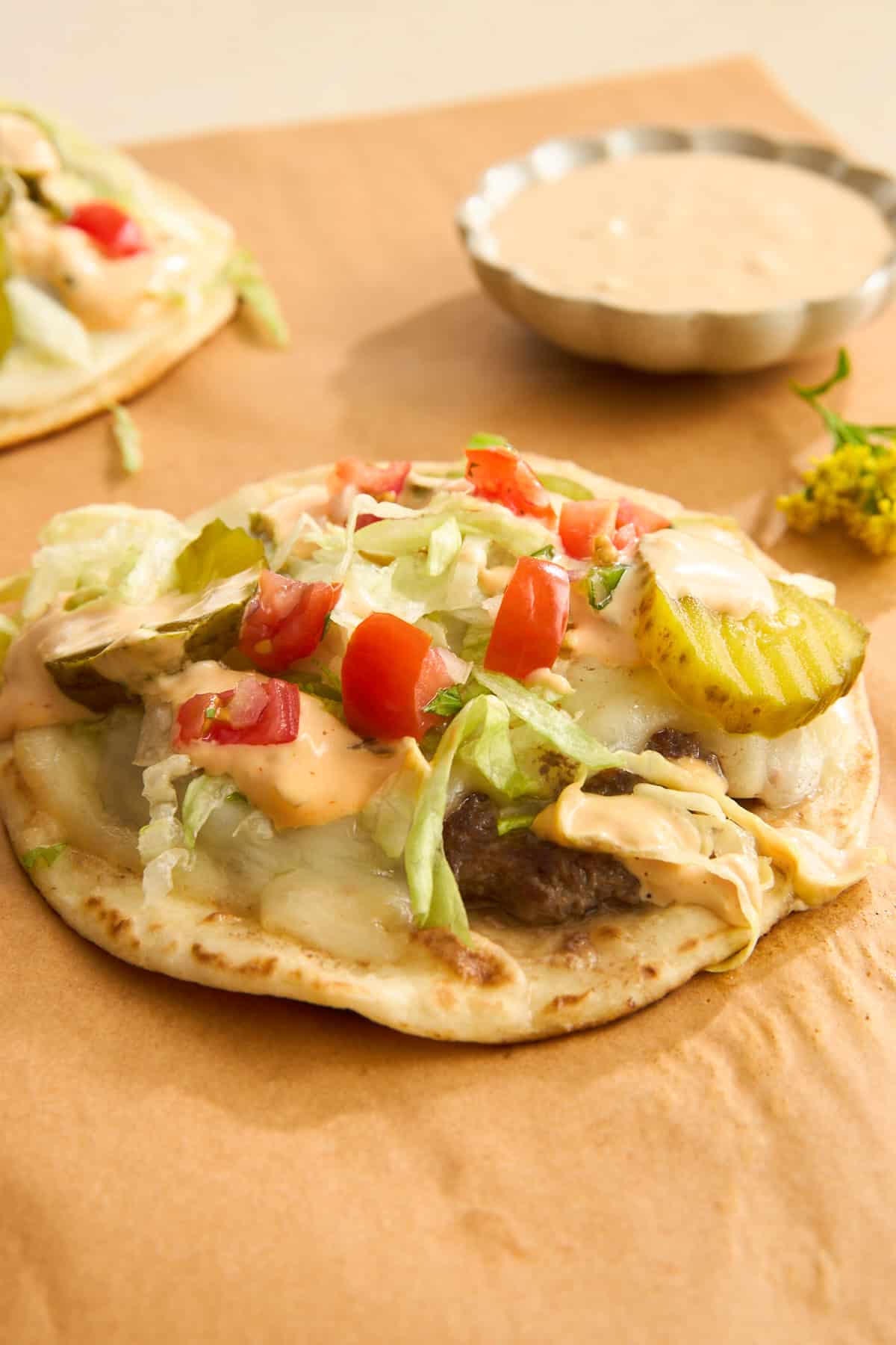A flatbread topped with ground beef, melted cheese, shredded lettuce, diced tomatoes, pickles, and a creamy sauce sits on brown parchment paper, with a small dish of extra sauce in the background.