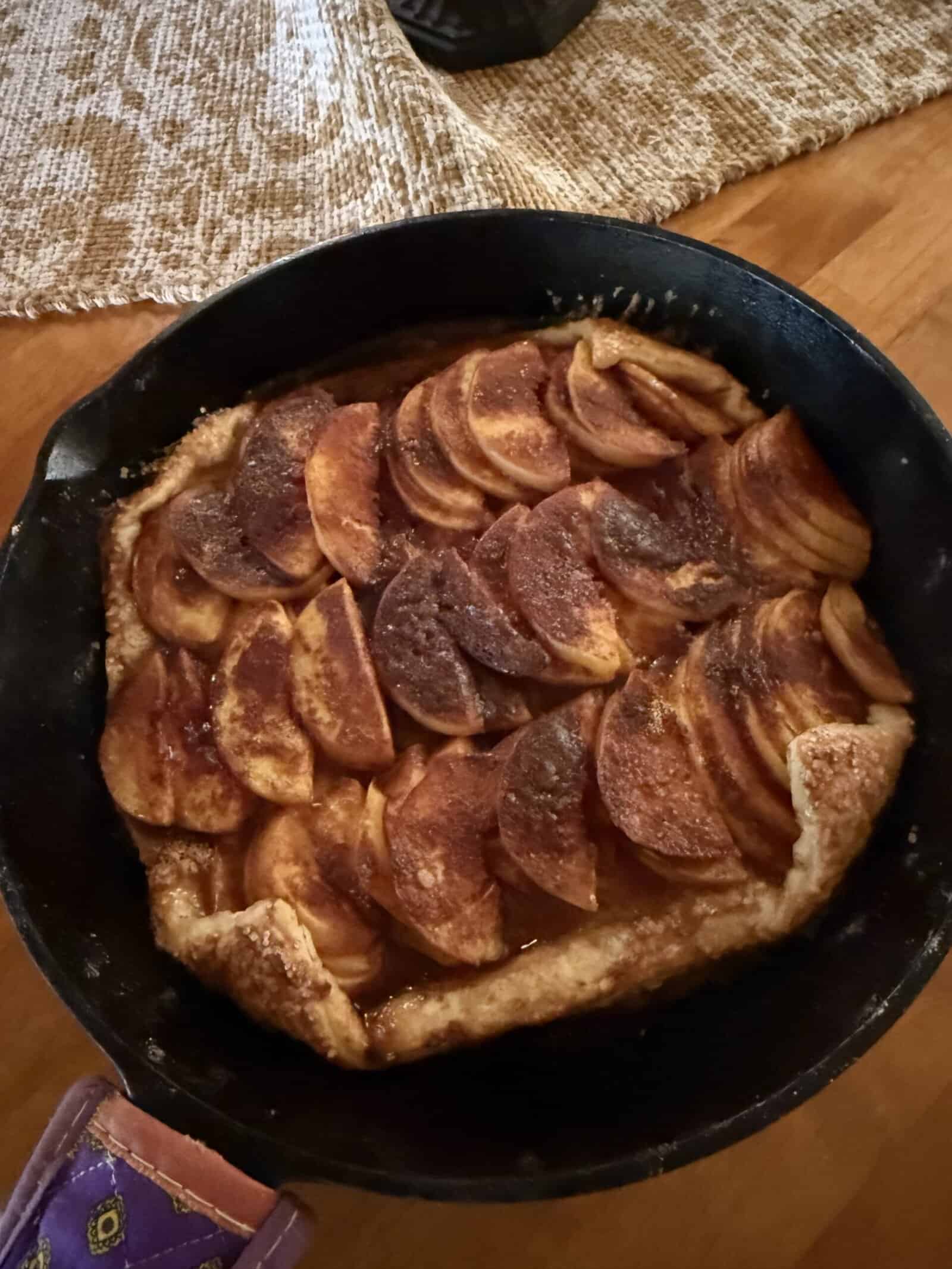 A rustic apple tart in a cast iron skillet, topped with neatly layered apple slices sprinkled with cinnamon and sugar, sits on a wooden table with a textured beige placemat in the background.