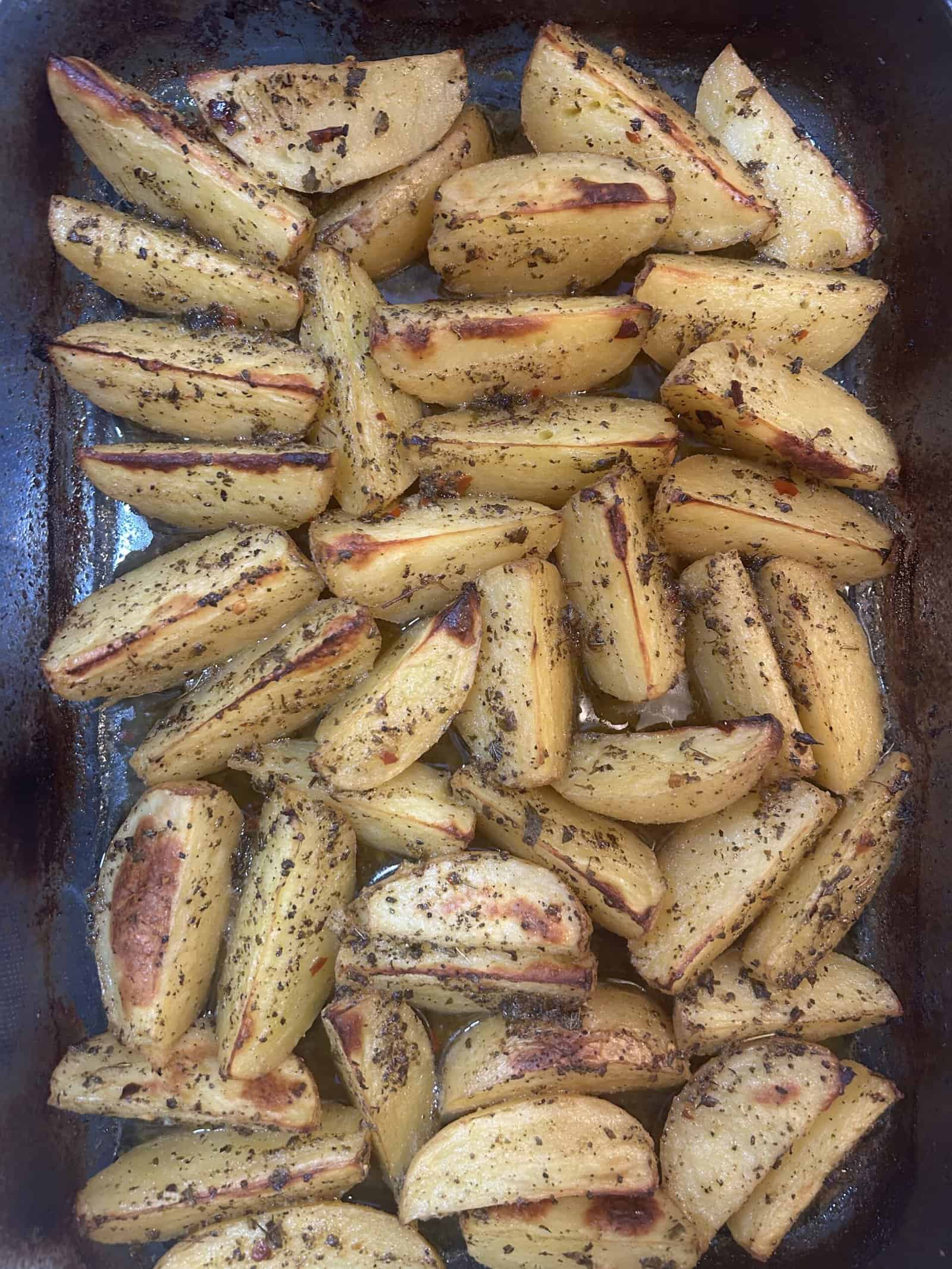 A baking tray filled with golden-brown potato wedges seasoned with herbs and spices, arranged in an even layer after roasting.