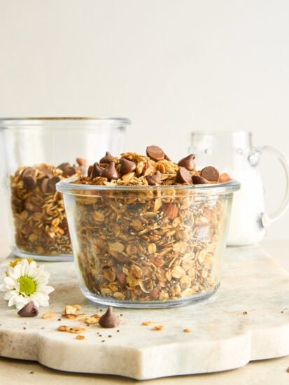 Two glass jars filled with granola and chocolate chips sit on a light stone surface. A small white flower and chocolate chips are scattered nearby. A glass pitcher of milk is in the background.