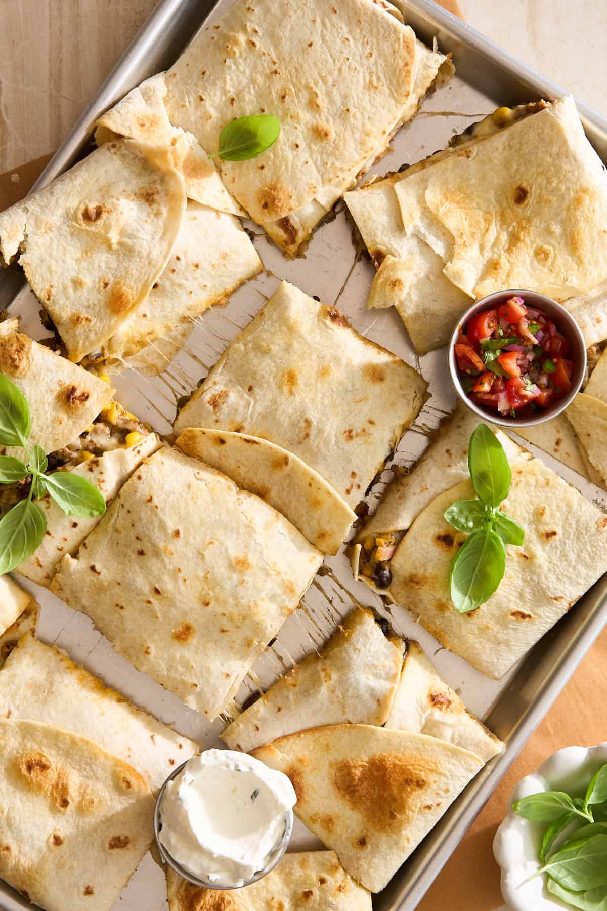 A tray of sliced quesadillas filled with cheese and vegetables, garnished with fresh basil leaves. A small bowl of salsa and a dish of sour cream are placed nearby for dipping.