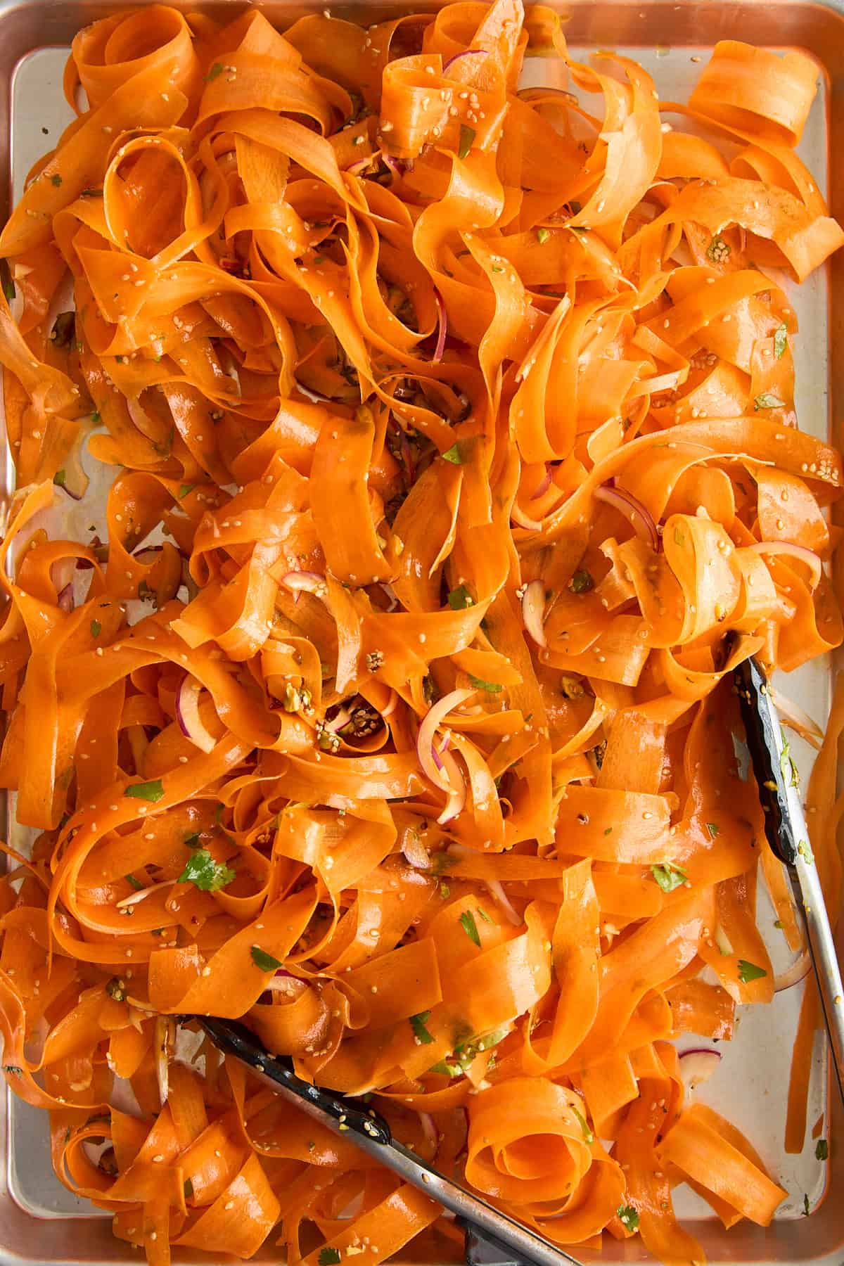 A tray filled with thin ribbons of carrot salad, garnished with herbs and seeds, with metal tongs resting on the side.