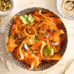 A bowl of carrot ribbon salad garnished with cilantro, red onion, sesame seeds, and pistachios sits on a light surface surrounded by a dish of sesame, a jar of chili flakes, and chopsticks.