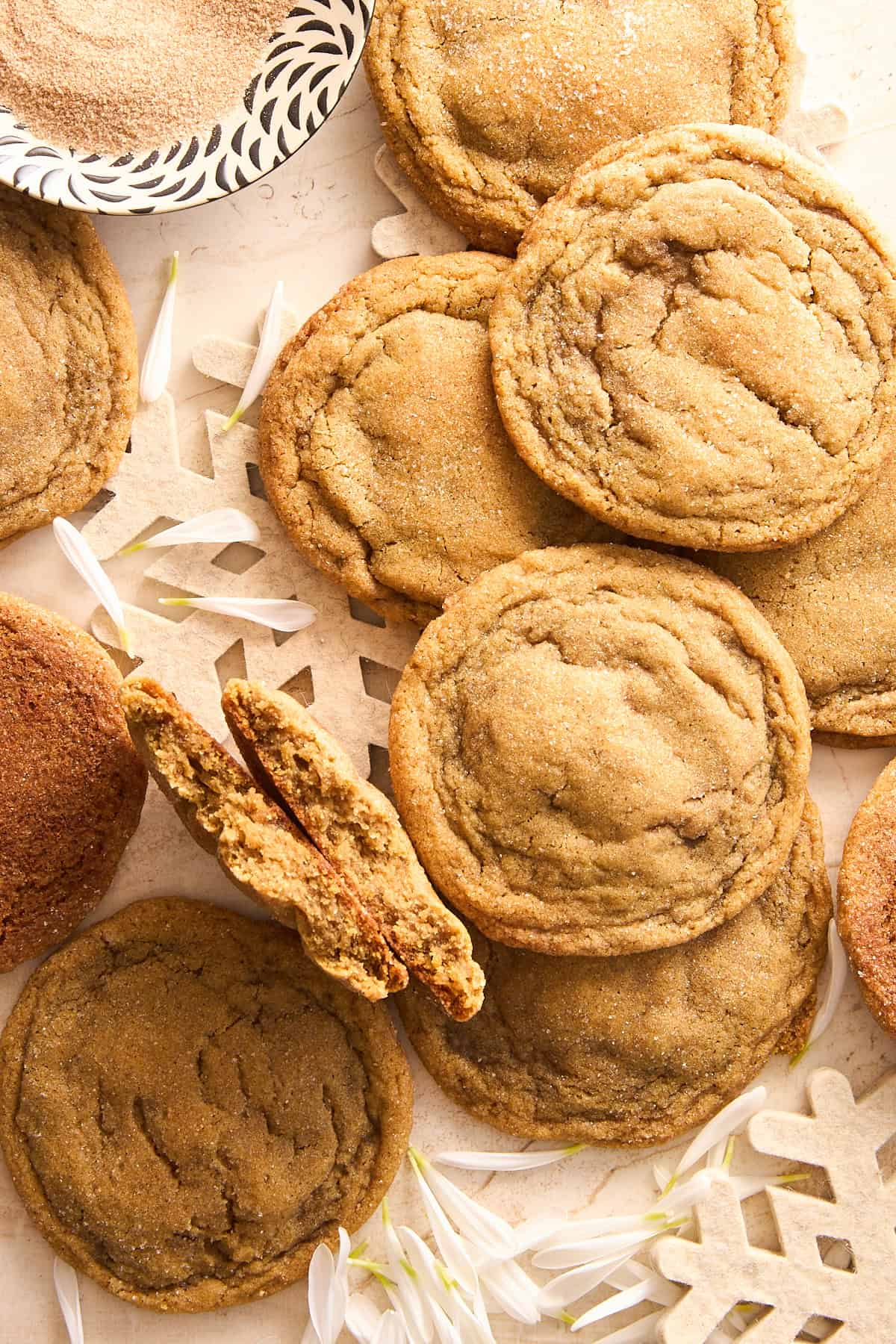 A close-up of several cinnamon sugar cookies on a light surface, with a few flower petals and a decorative paper snowflake. One cookie is broken in half, showing its soft interior. A bowl of cinnamon sugar is in the corner.