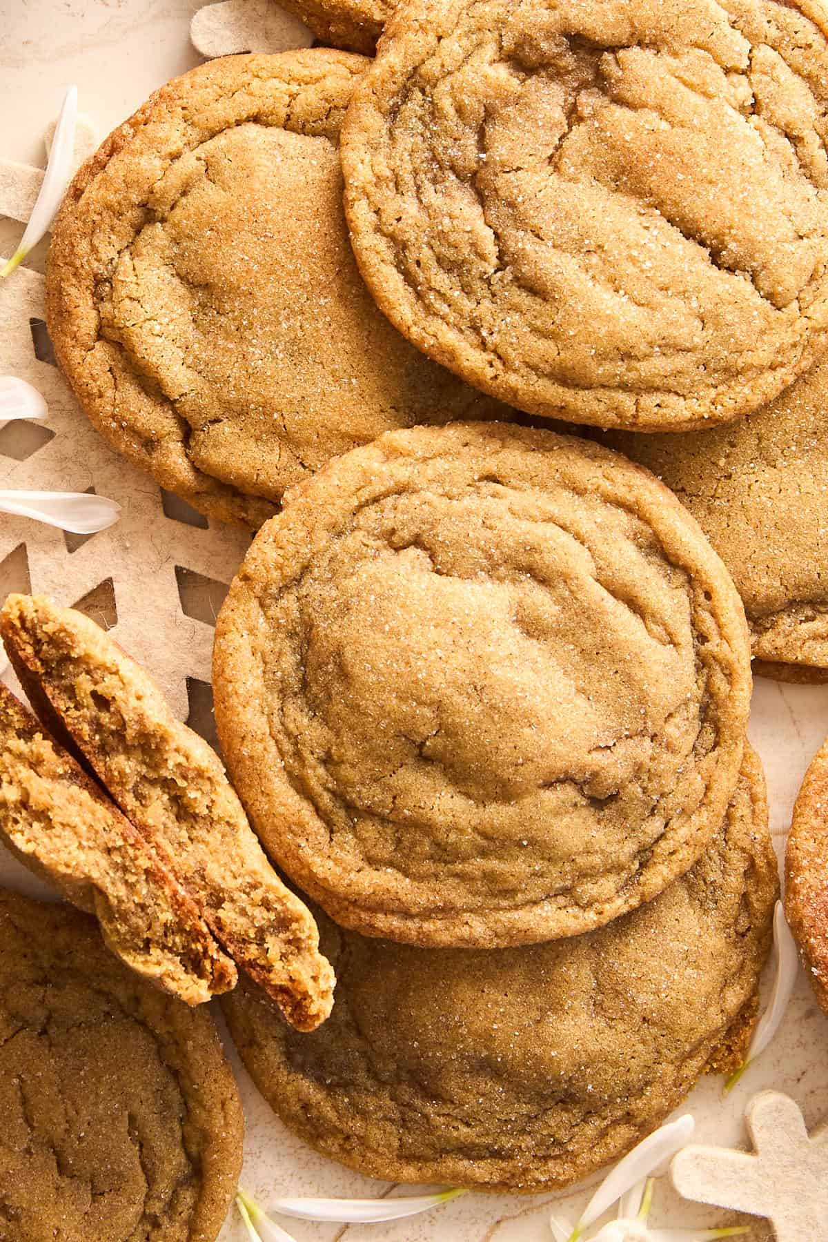 A close-up of several golden brown cookies, some stacked and one cookie broken in half, revealing a soft, chewy interior. The cookies have a slightly crackled surface and are placed on a light-colored surface.
