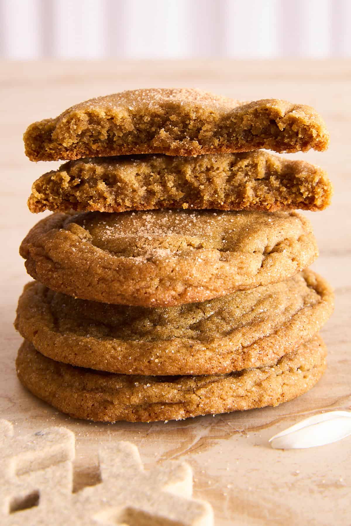 A stack of five brown sugar cookies, with the top cookie broken in half to show a soft, chewy texture inside. The cookies are sprinkled with sugar and rest on a light wooden surface.