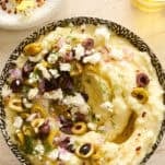 A bowl of creamy mashed potatoes topped with chopped olives, crumbled feta cheese, diced red onions, and fresh dill, with a drizzle of olive oil. A small dish of red pepper flakes and a jar of olive oil are nearby.