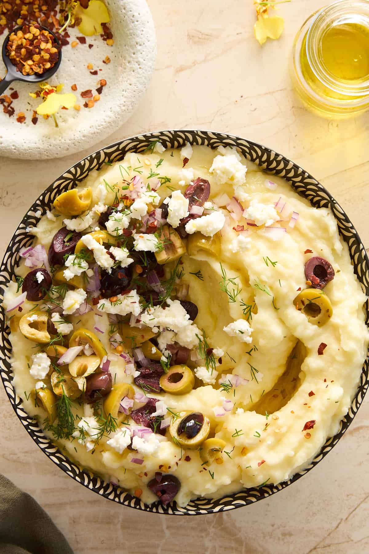 A bowl of creamy mashed potatoes topped with chopped olives, crumbled feta cheese, diced red onions, and fresh dill, with a drizzle of olive oil. A small dish of red pepper flakes and a jar of olive oil are nearby.