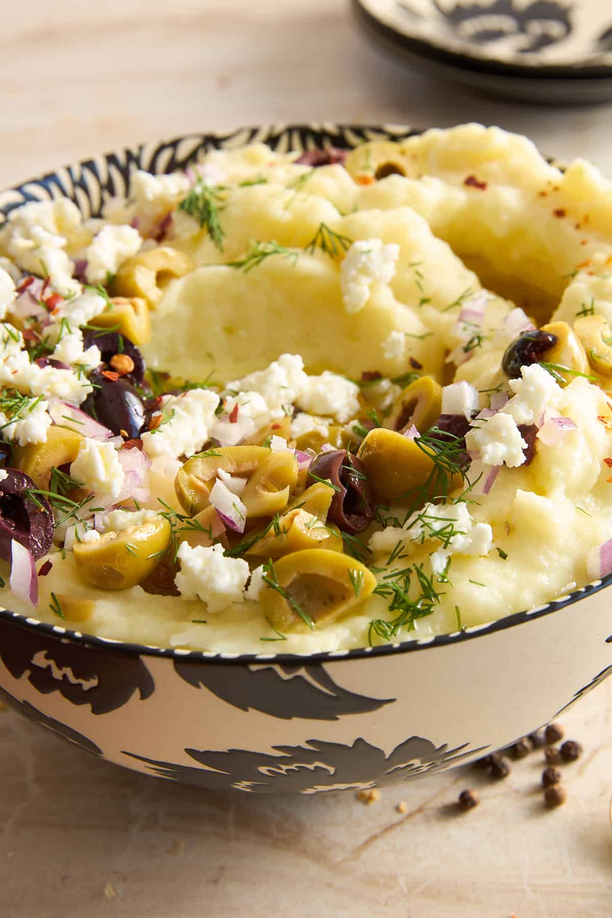 A bowl of mashed potatoes topped with chopped green and black olives, crumbled feta cheese, red onion, dill, and red pepper flakes, in a decorative black and white bowl.