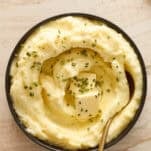 A bowl of creamy mashed potatoes topped with melting butter and chopped chives, with a gold spoon resting inside. The bowl sits on a light-colored marble surface.