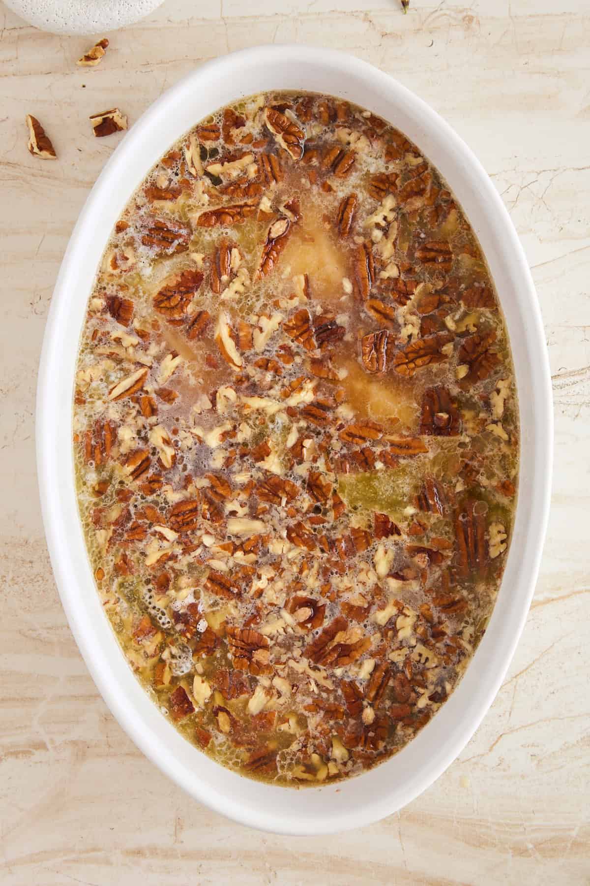 An oval white baking dish filled with a liquid mixture topped with chopped pecans, ready to be baked, sits on a light marble surface.