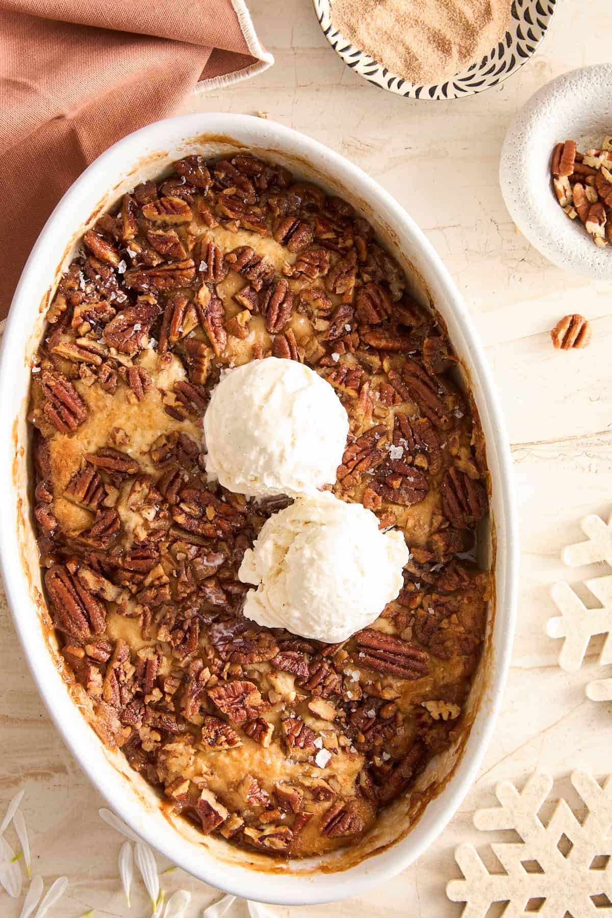 An oval dish filled with pecan bread pudding topped with two scoops of vanilla ice cream, surrounded by a napkin, a bowl of pecans, and snowflake-shaped decorations on a beige surface.