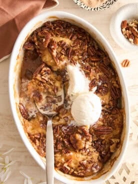 An oval baking dish filled with pecan cobbler, topped with two scoops of vanilla ice cream. A serving spoon rests in the dish, and some cobbler has been served. Pecan pieces and a brown napkin are nearby.