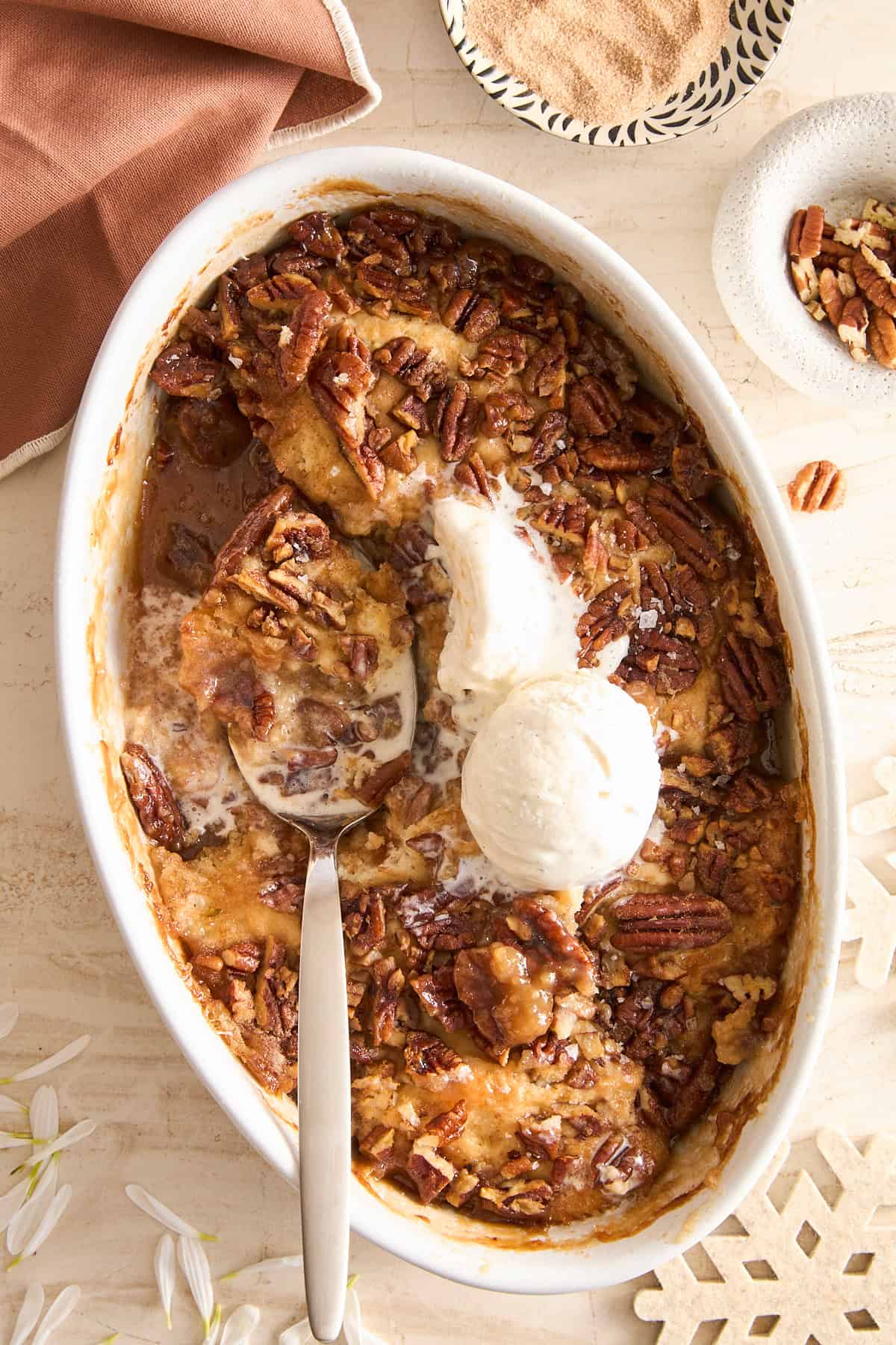 An oval baking dish filled with pecan cobbler, topped with two scoops of vanilla ice cream. A serving spoon rests in the dish, and some cobbler has been served. Pecan pieces and a brown napkin are nearby.
