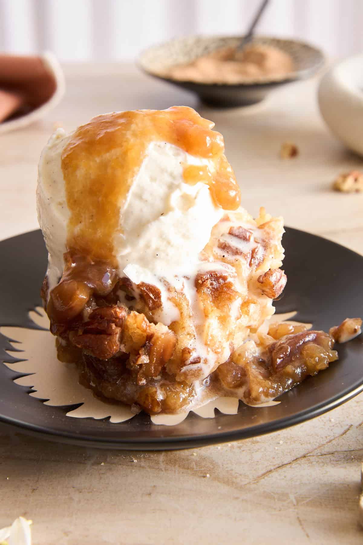 A slice of pecan cobbler topped with a scoop of vanilla ice cream and caramel sauce sits on a black plate, with melted ice cream dripping down the sides. A bowl and spoon are blurred in the background.