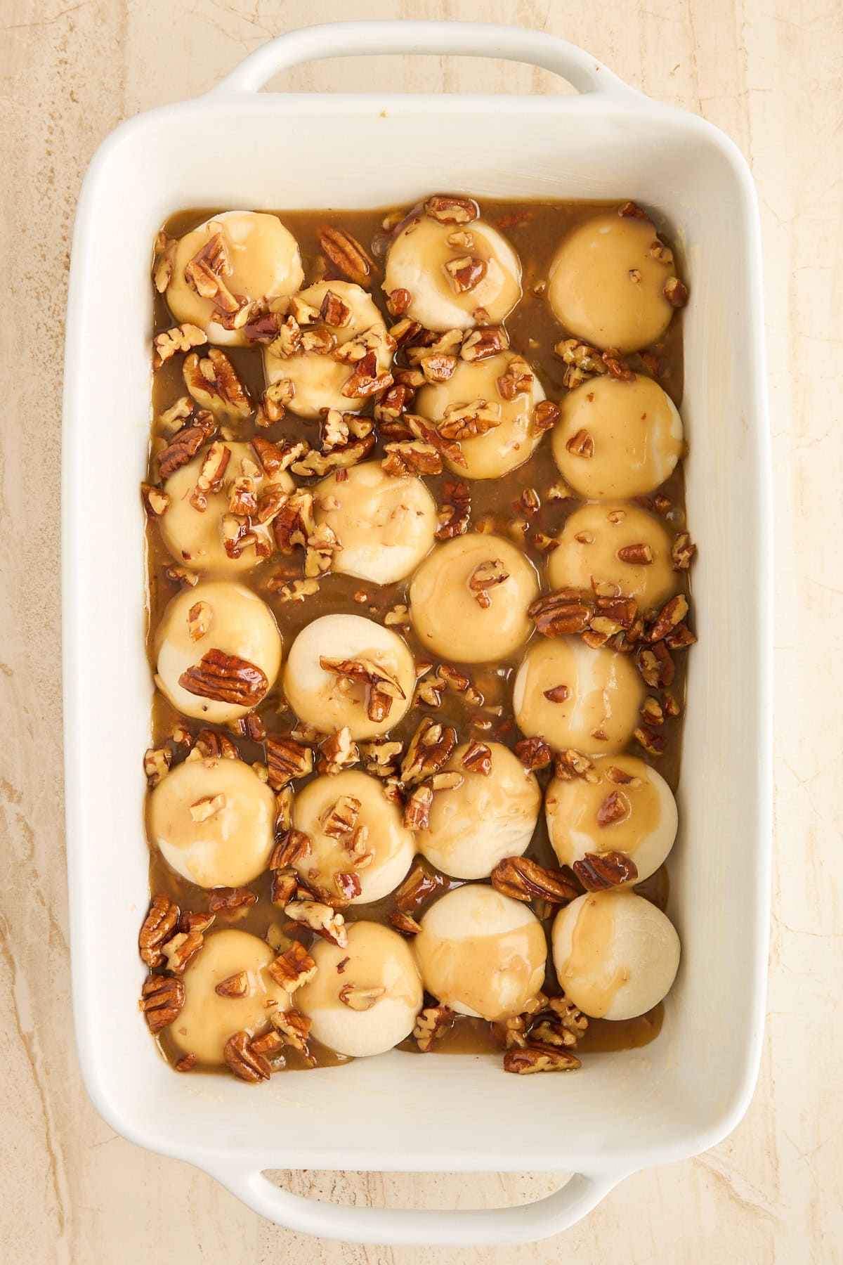 A white baking dish filled with round dough balls topped with brown caramel sauce and chopped pecans, ready to be baked. The dish is placed on a light-colored surface.