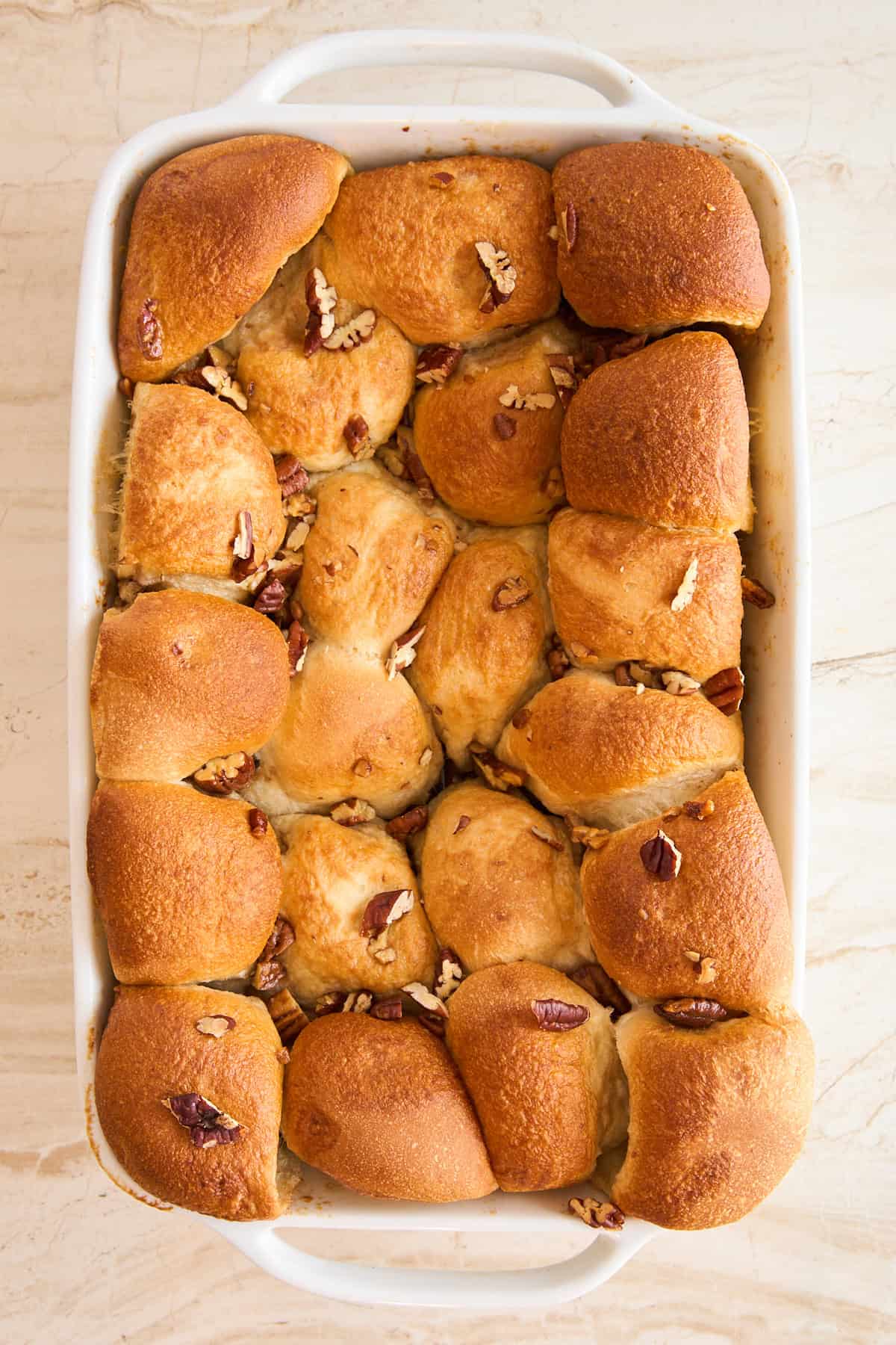 A white baking dish filled with golden-brown pull-apart rolls, topped with chopped pecans, on a light-colored surface.