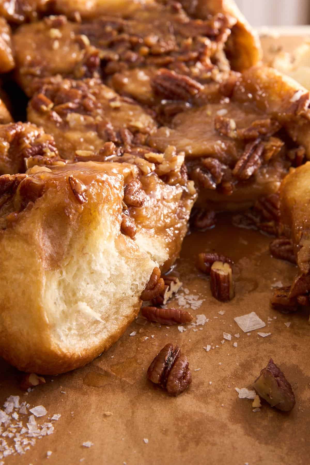 Close-up of sticky pecan buns covered in caramel sauce and chopped pecans, resting on brown parchment paper with a few granules of flaky sea salt scattered around.