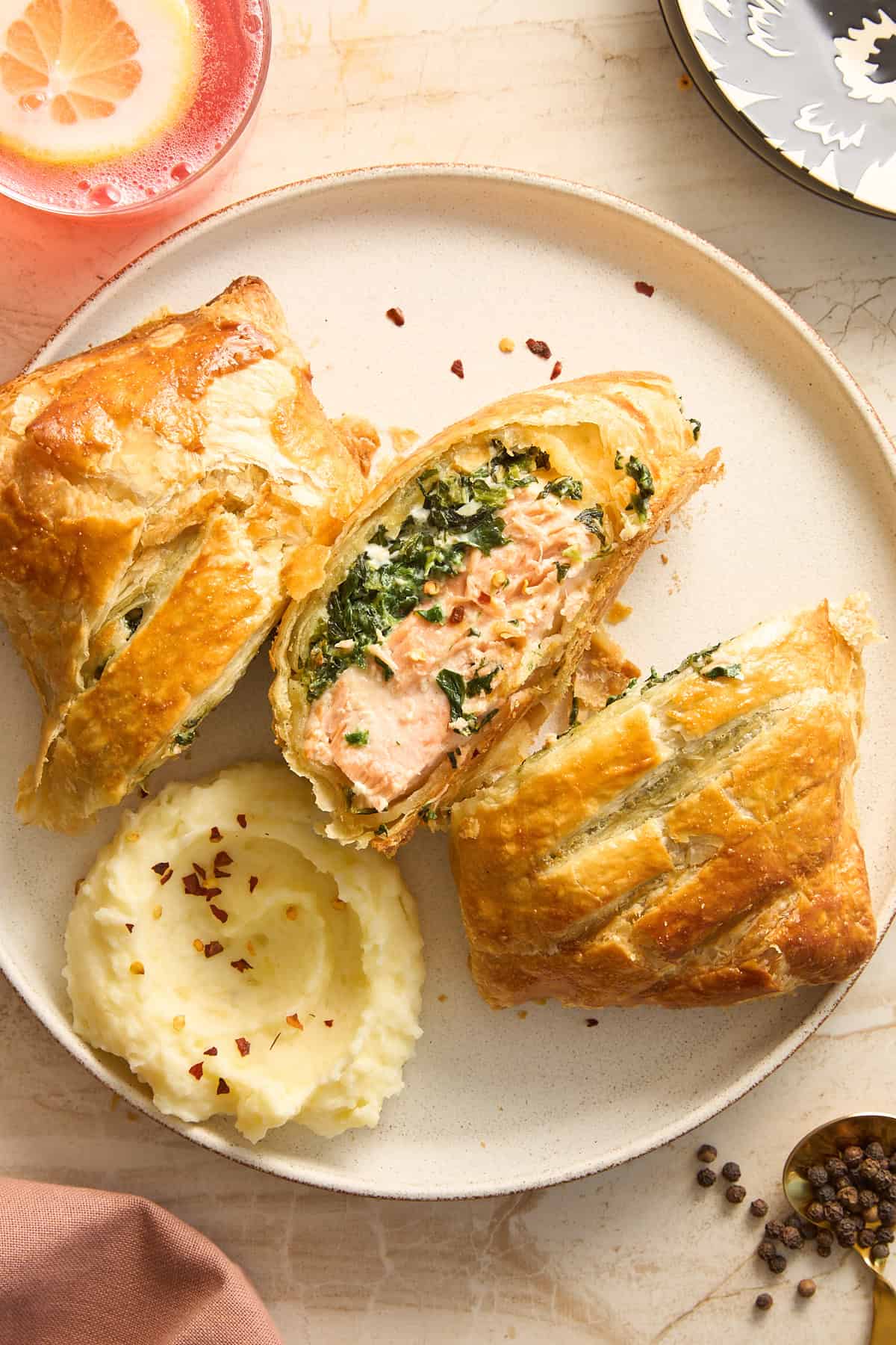 A plate with salmon Wellington sliced open to reveal spinach filling, served with mashed potatoes sprinkled with red pepper flakes. A pink drink with lemon and peppercorns are nearby on a marble surface.