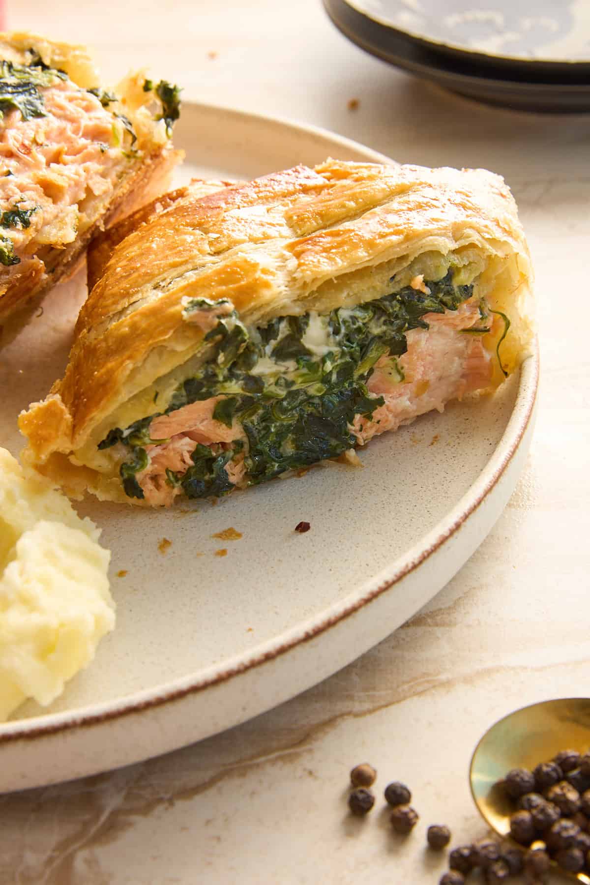 A close-up of a slice of salmon Wellington with spinach and pastry crust on a beige plate, served with mashed potatoes. Black peppercorns and a spoon are nearby on a light-colored surface.