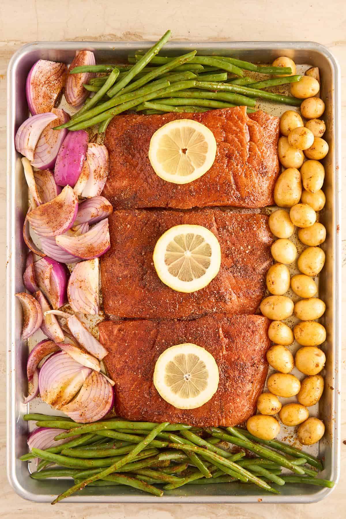A sheet pan with three seasoned salmon fillets, each topped with a lemon slice, surrounded by green beans, halved red onions, and small golden potatoes, all arranged neatly before roasting.