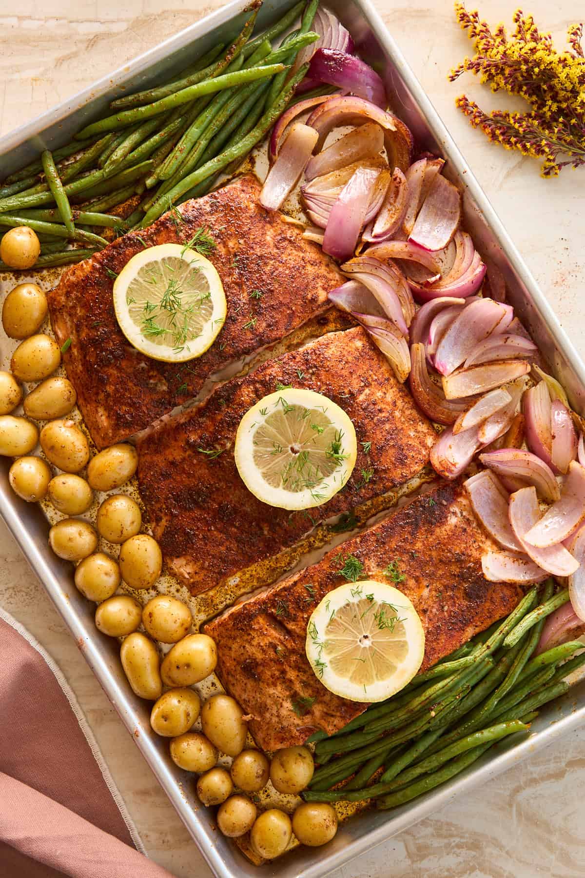 A sheet pan with three seasoned salmon fillets topped with lemon slices and fresh herbs, surrounded by green beans, roasted red onions, and baby potatoes.