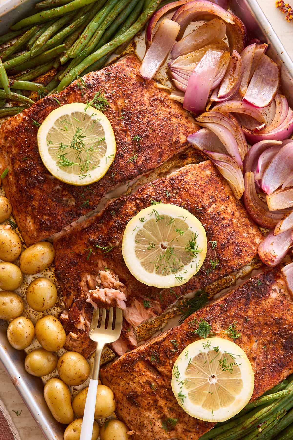 A sheet pan with seasoned salmon fillets topped with lemon slices and dill, surrounded by roasted green beans, red onion wedges, and small yellow potatoes; a fork flakes a piece of the salmon.