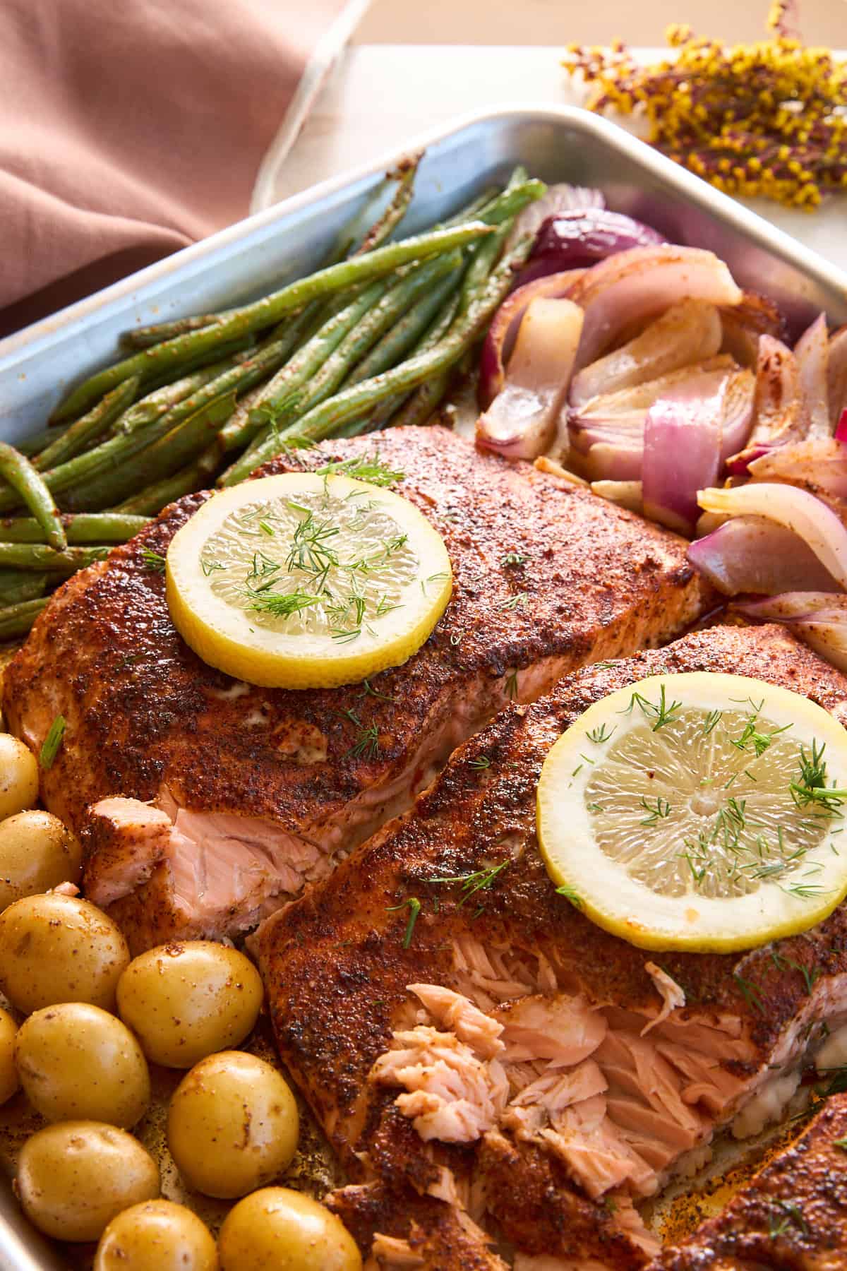 A baking tray with seasoned salmon fillets topped with lemon slices, roasted baby potatoes, green beans, and red onions. The food is garnished with fresh herbs and arranged neatly for serving.