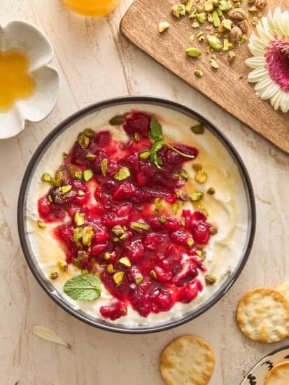 A bowl of creamy dip topped with red fruit sauce, chopped pistachios, and mint leaves sits on a marble surface. Nearby are crackers, a small dish of honey, orange juice, and a wooden board with chopped pistachios and a pink flower.