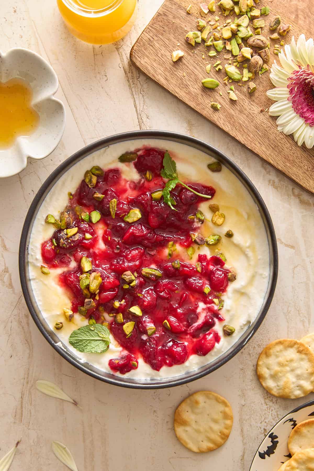 A bowl of creamy whipped feta dip topped with red fruit sauce, chopped pistachios, and mint leaves sits on a marble surface. Nearby are crackers, a small dish of honey, orange juice, and a wooden board with chopped pistachios and a pink flower.