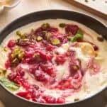 A bowl of creamy yogurt topped with swirling raspberry sauce, chopped pistachios, and fresh mint leaves sits on a light surface, with honey and more pistachios visible in the background.