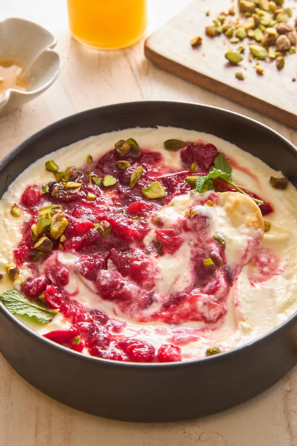A bowl of creamy yogurt topped with swirling raspberry sauce, chopped pistachios, and fresh mint leaves sits on a light surface, with honey and more pistachios visible in the background.