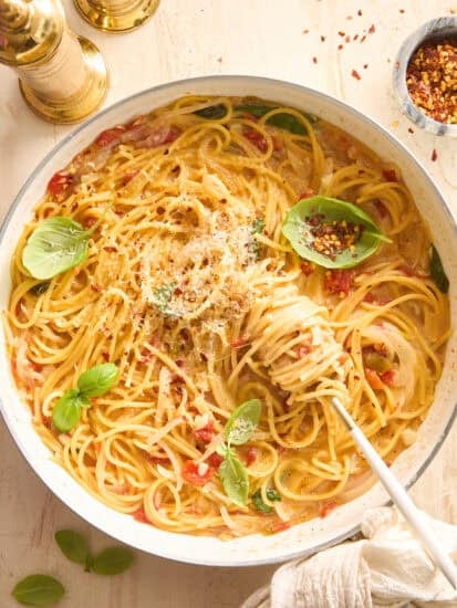 A skillet filled with creamy spaghetti garnished with fresh basil and grated cheese. A fork twirls some pasta, and there are gold salt and pepper shakers, red pepper flakes, and basil leaves nearby on a light table.