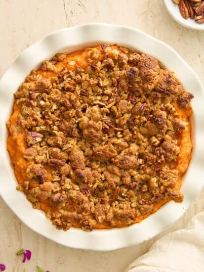 A round white dish filled with sweet potato casserole, topped with a golden-brown pecan and oat crumble. The dish sits on a light-colored surface, with a bowl of pecans and flower petals nearby.