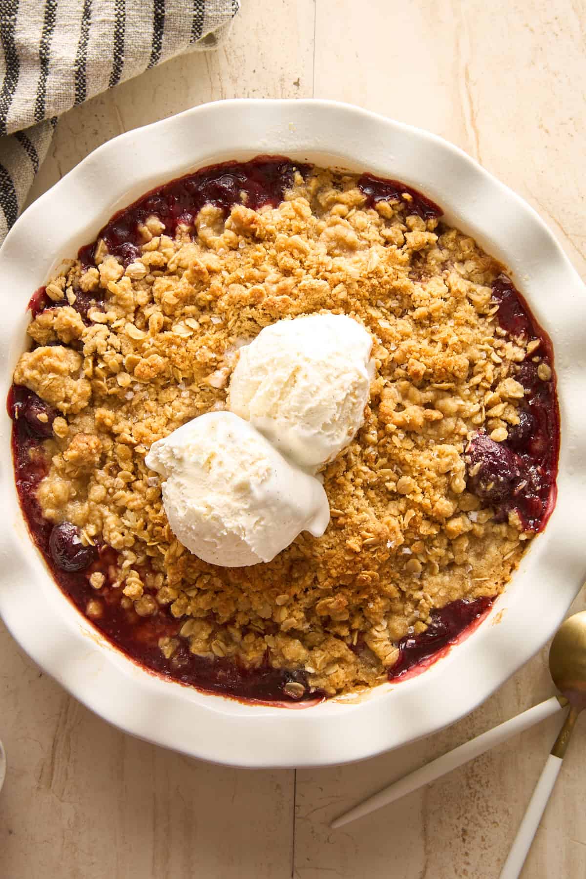 A baked cherry crisp topped with a golden oat crumble and two scoops of vanilla ice cream, served in a white ceramic pie dish on a light surface with a striped towel and gold spoon nearby.