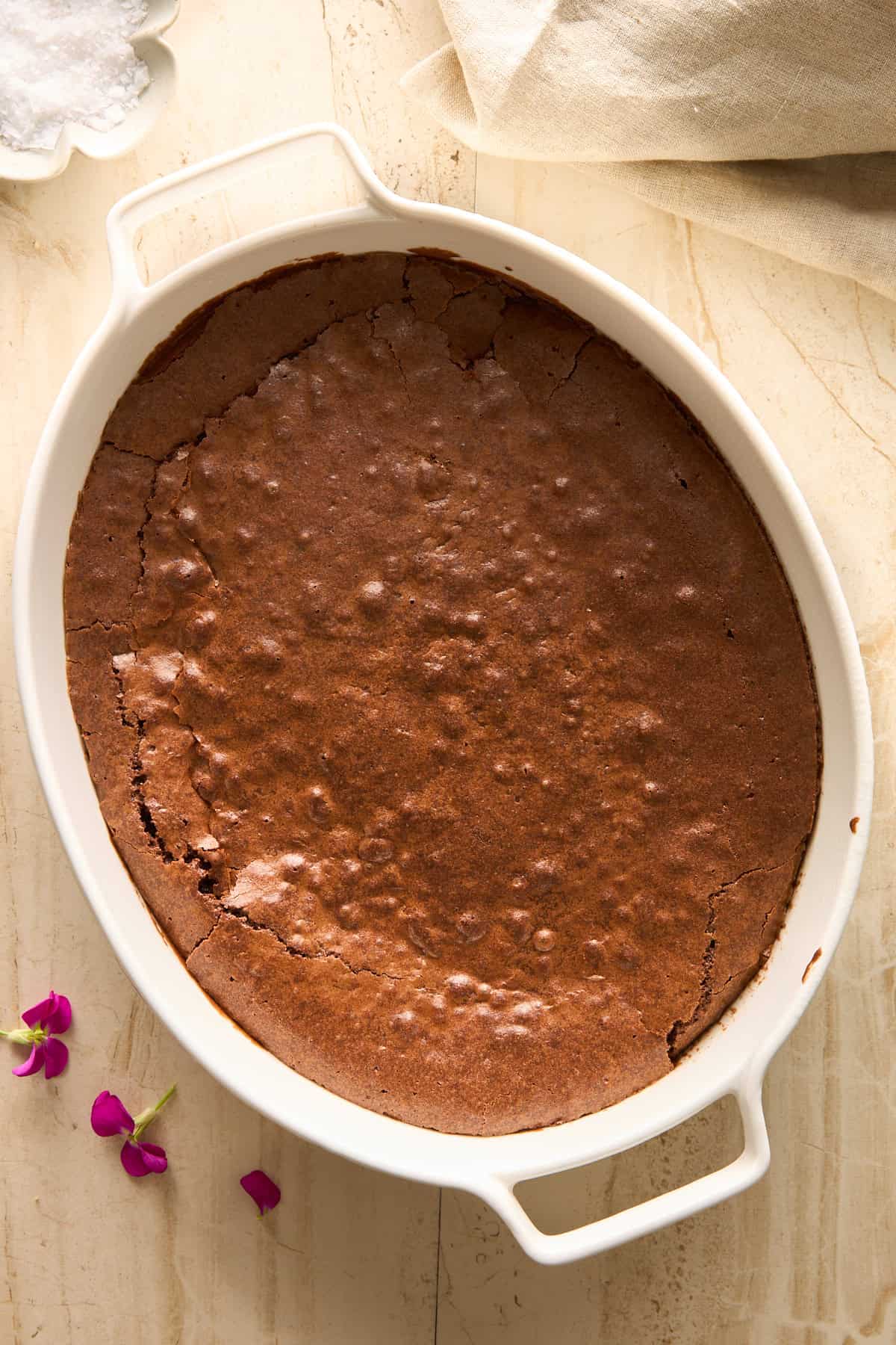 An oval white baking dish filled with a freshly baked chocolate cake, with a crackled surface, sits on a light countertop. A few pink flower petals and a bowl of salt are nearby.