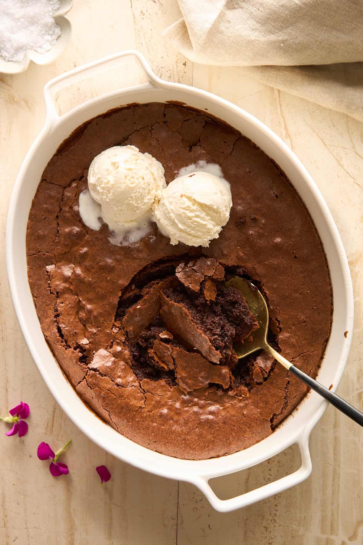 A white baking dish with chocolate brownie and two scoops of vanilla ice cream on top. A spoon has taken a serving out, showing the gooey brownie inside. Pink flowers and a beige cloth are nearby on a light surface.