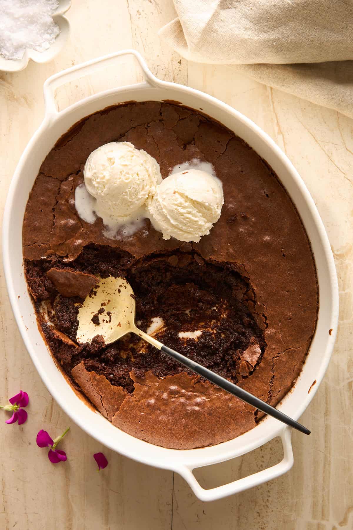 An oval white baking dish holds a chocolate cake with a large scoop missing, revealing a gooey interior. Two scoops of vanilla ice cream sit on top, with a serving spoon resting inside. Pink flowers are beside the dish.
