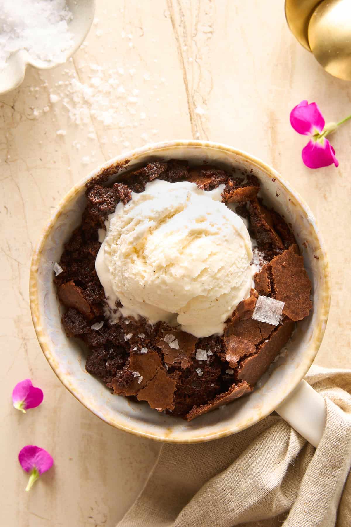 A bowl of chocolate brownie topped with a scoop of vanilla ice cream and sea salt flakes, surrounded by pink flower petals on a light-colored surface.